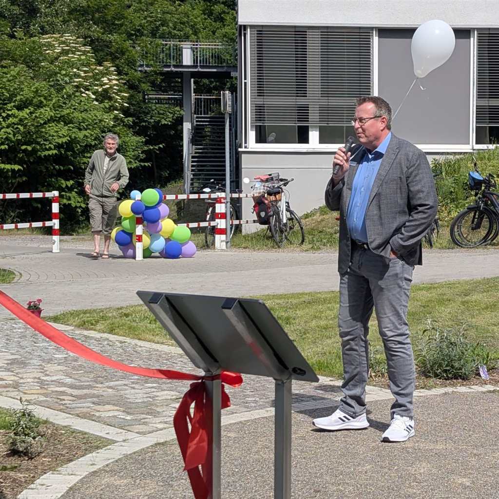 Ein Mann in einem grauen Anzug hält ein Mikrofon und einen weißen Ballon und spricht vor einem Gebäude. Im Hintergrund stehen zwei Männer, ein Haufen 