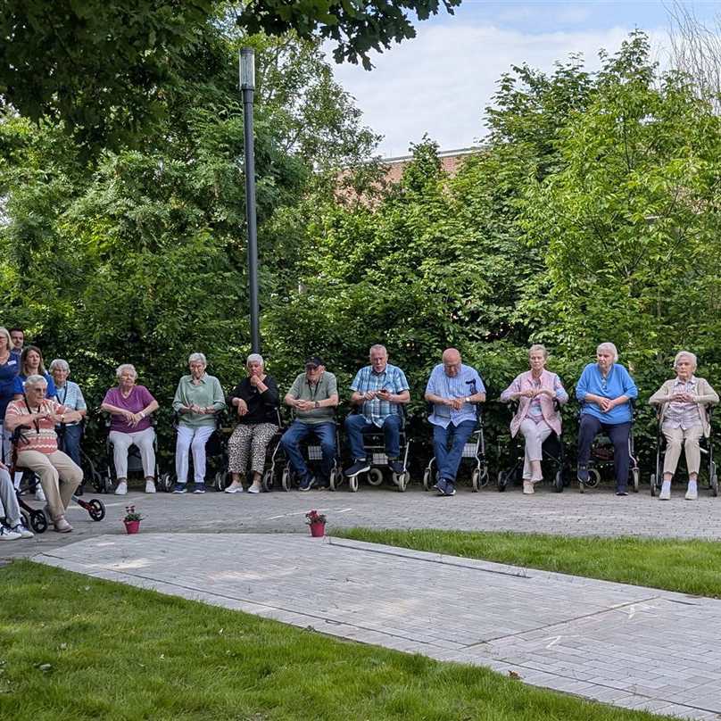 Senioren sitzen nebeneinander auf Stühlen und Rollstühlen im Freien, einige blicken in die Kamera.
