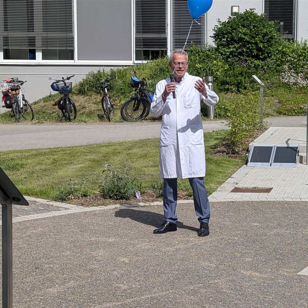 Mann in Arztkittel hält eine Ansprache vor einem Gebäude