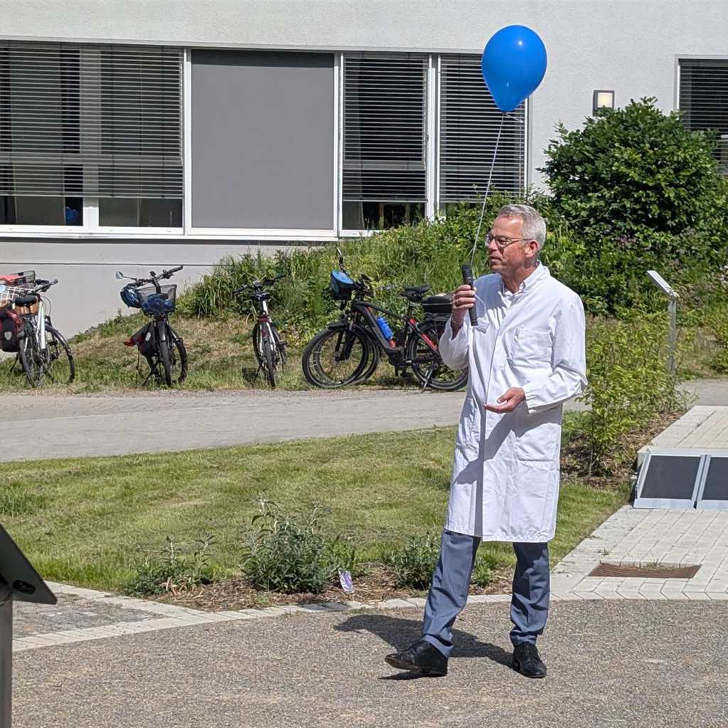 Ein Mann im weißen Kittel hält ein Mikrofon und einen blauen Ballon in der Hand.