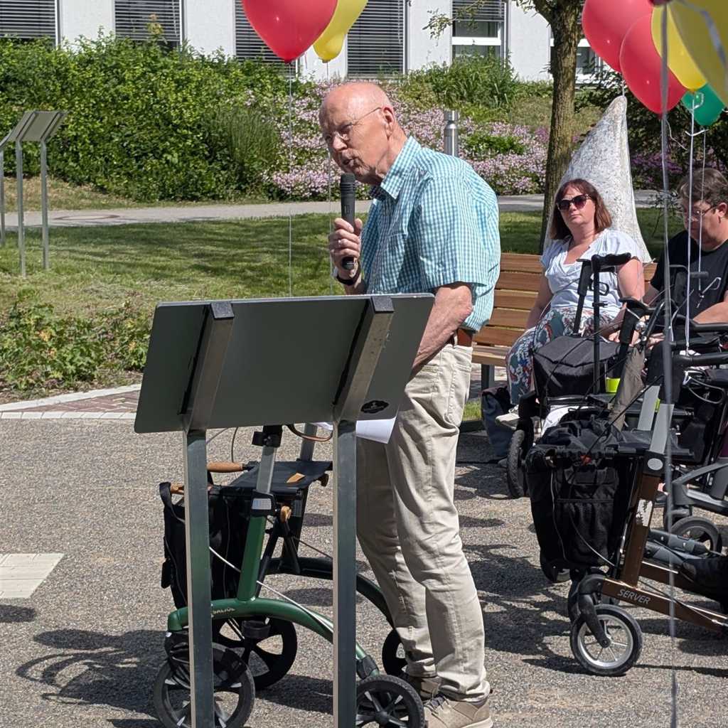 Ein älterer Mann mit Mikrofon spricht zu einem Publikum von Rollator-Nutzern im Freien.