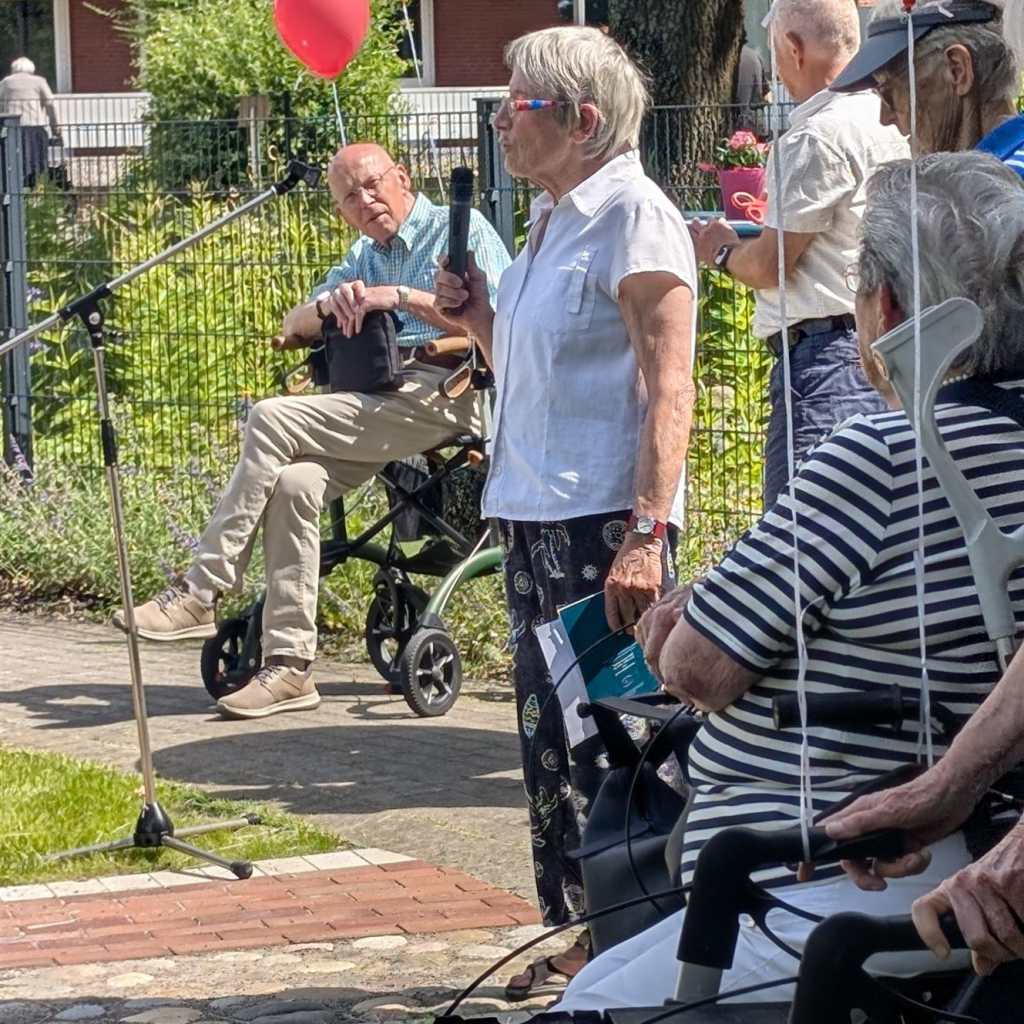 Frau hält Mikrofon und spricht, neben ihr sitzt ein älterer Mann im Rollstuhl, im Hintergrund sind Ballons und weitere Personen zu sehen.